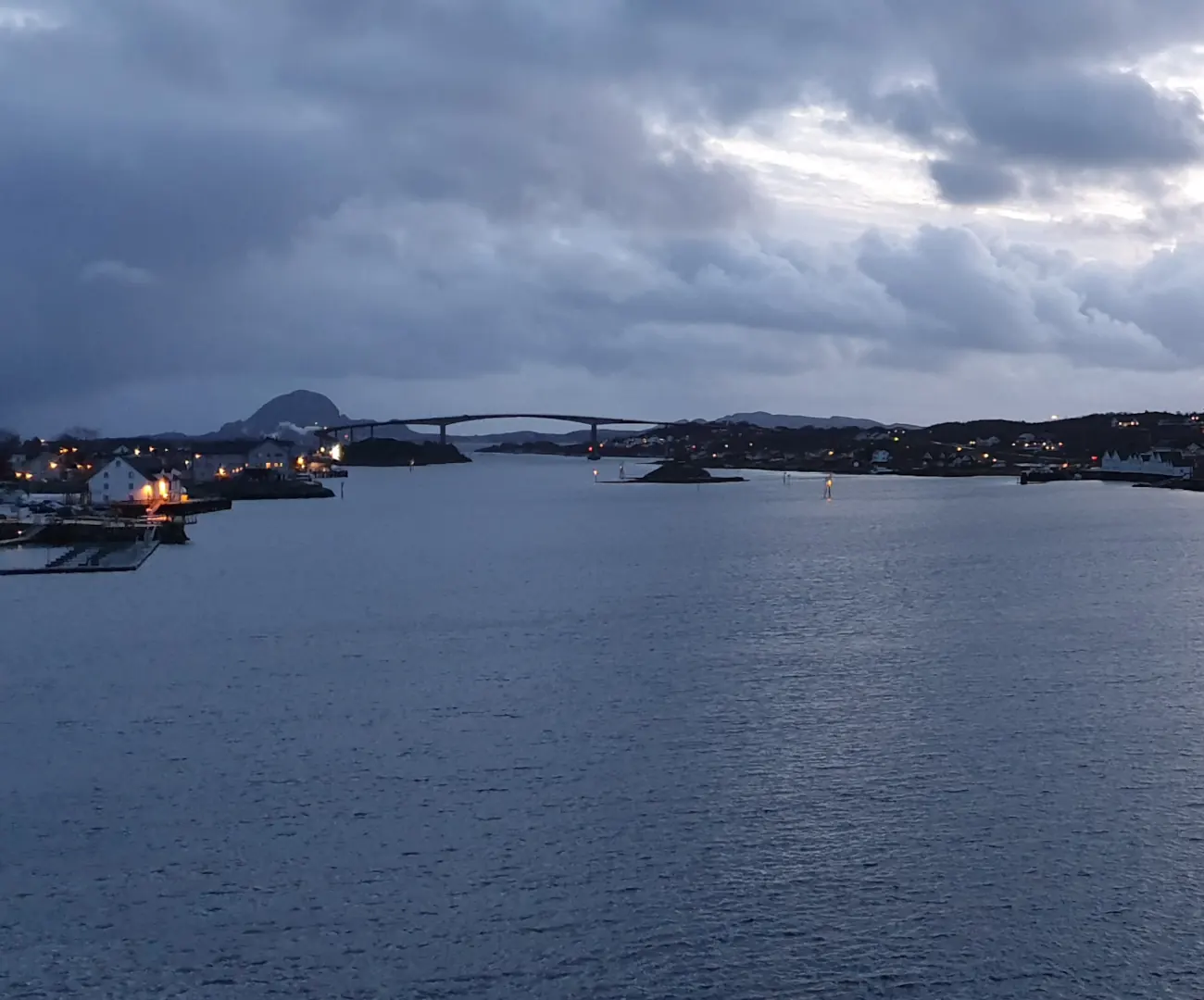 Brønnøysund i skumringen sett fra sjøen, med lys i husene langs sundet, Brønnøysundbrua og Torghatten i bakgrunnen under tunge skyer