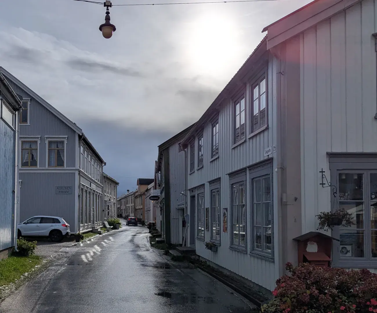 Sjøgato i Mosjøen etter en regnskur, med gamle trehus langs en blank, våt gate og sola som bryter gjennom skyene