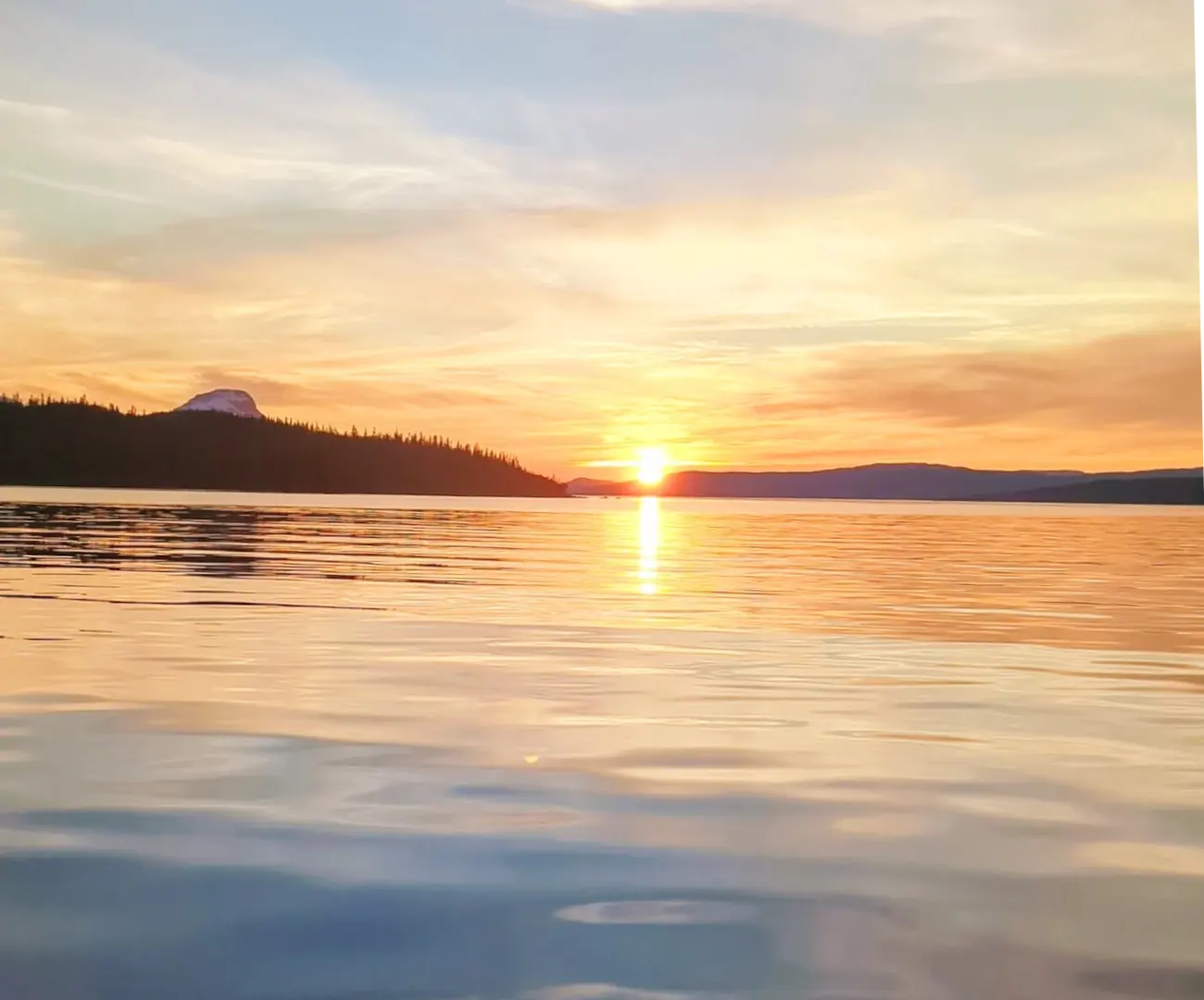 Solnedgang over Røssvatnet, med skogkledde åser og en snødekt fjelltopp i silhuett og sola speilet i det blanke vannet