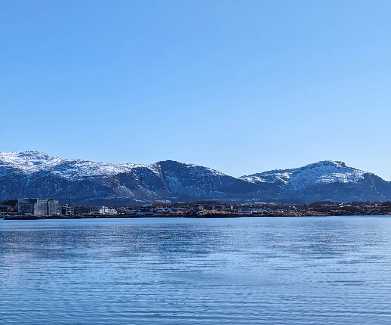 Sandnessjøen sett fra sjøen en klar vårdag, med byen langs vannkanten og snødekte fjell bak under en blå himmel