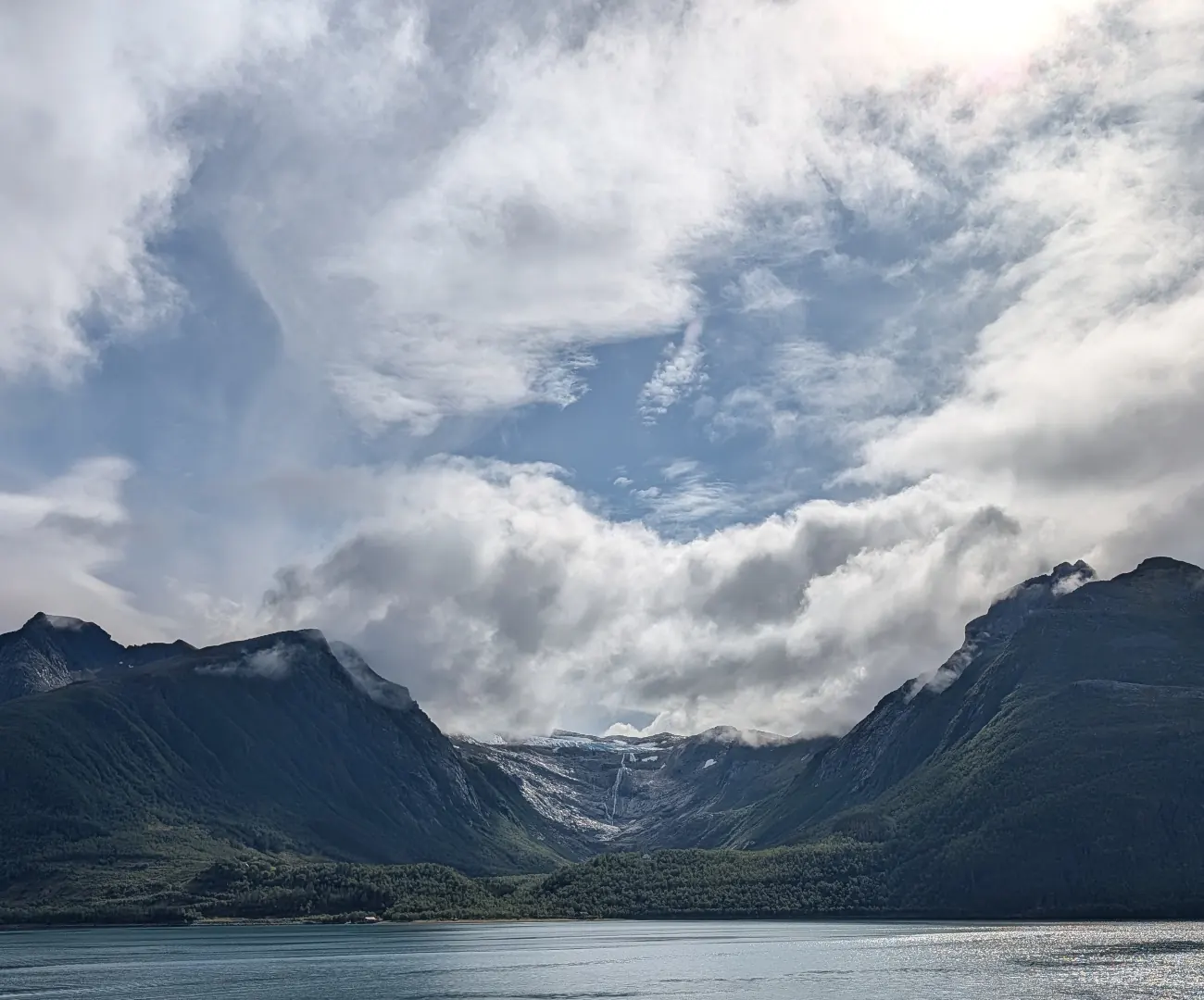 Svartisen sett fra fjorden, med en isbre og et smalt fossefall mellom mørke fjell under store, hvite skyer