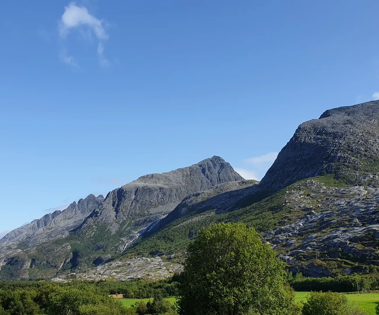 De syv søstre ved Sandnessjøen en solskinnsdag, med bratte, grå fjelltopper over grønne lier og en lys blå himmel