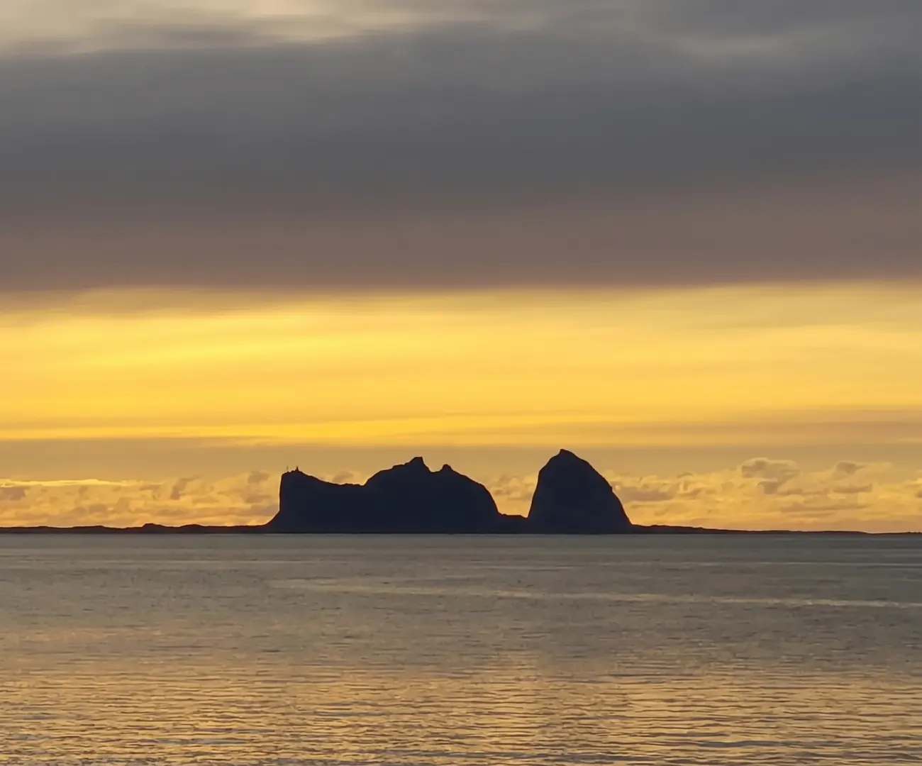 Fjellene på Træna som mørk silhuett mot en gul solnedgang over et rolig hav